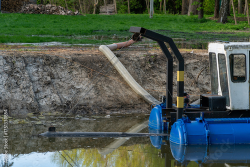 Industrial dredging machinery floating on a small body of water near an exposed earthen slope with green foliage