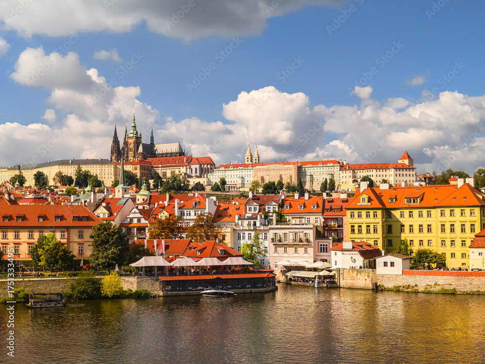 Obraz premium Beautiful landscape of a historical riverside city area with vibrant yellow buildings and red roofs reflecting in the river water under a bright sky with large white cumulus clouds.