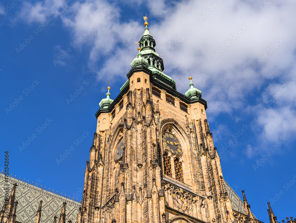 Obraz premium Close up of a magnificent cathedral clock tower with green spires and golden details reaching into a vibrant blue sky with soft white clouds.