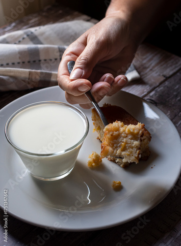 Manipulacion de alimentos, manejo de comida en la cocina, mano con cuchara partiendo un pastel o pan de elote sobre un plato blanco con un vaso de leche, vertical.