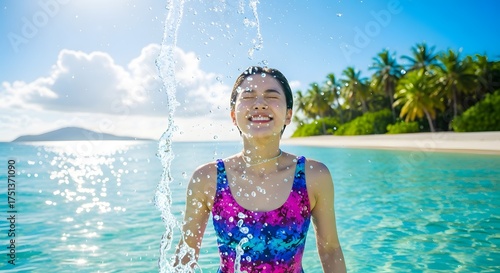 Joyful young woman splashes water in turquoise ocean, eyes closed, smiling blissfully amidst a tropical paradise beach scene with palm trees.