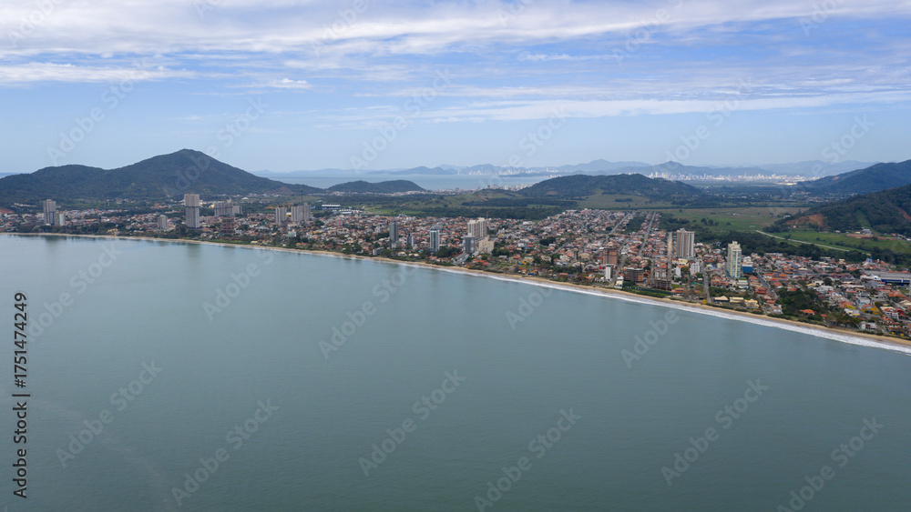 Fototapeta premium Aerial frontal view of Penha Beach with calm sea, coastal cityscape, and mountains in the background, Santa Catarina, Brazil.