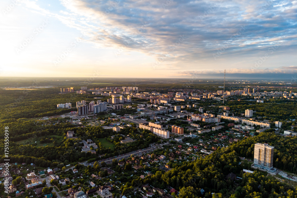 Obraz premium Aerial view of a city immersed in lush green forests at golden hour. Urban landscape with buildings, houses, and roads under a cloudy sky