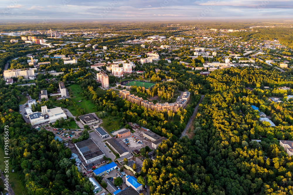 Obraz premium Aerial view of a green city with modern residential buildings, a stadium, and vast forests under a golden hour sky. Urban landscape surrounded by nature