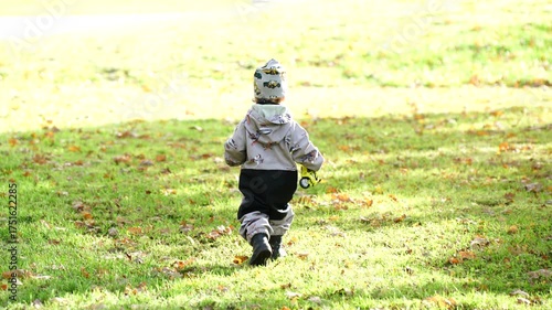 Rear view of small child wearing warm outdoor clothing walking alone on grassy park field in autumn sunlight showing childhood exploration, independence and healthy outdoor lifestyle