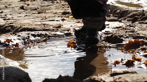 Close view of child’s waterproof boots stepping into muddy puddle with fallen autumn leaves showing realistic ground texture, wet surface reflection and outdoor play environment