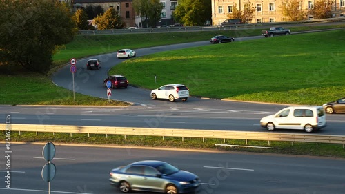Aerial view of several cars moving through curved road interchange surrounded by green grass and city buildings in evening light showing organized traffic flow and urban transport system