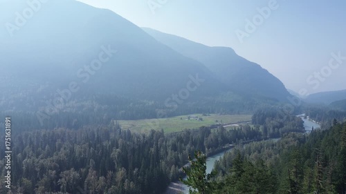 Cinematic drone footage captures the serene beauty of a remote wilderness. The camera glides over pine treetops to showcase the winding river and misty, layered mountain peaks.