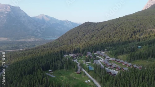 This cinematic drone footage captures a charming mountain town from a high angle. The buildings are surrounded by green trees, with a majestic mountain range visible in the distance.