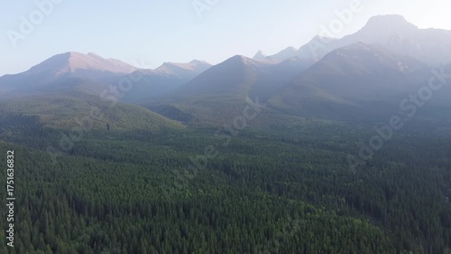 A cinematic drone flight over a remote mountain wilderness at dawn. The rising sun creates a beautiful, atmospheric haze that settles between the majestic, layered peaks.