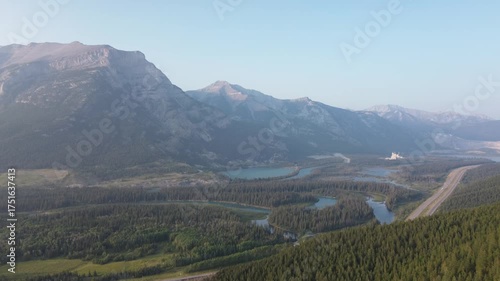 Cinematic drone flight over a remote mountain pass. The road winds through a pristine forest landscape, leading towards majestic, sun-kissed peaks in the distance.