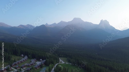 Cinematic drone flight over a remote mountain village at dawn. The community is surrounded by a dense pine forest, with majestic, sun-kissed peaks creating a breathtaking backdrop.