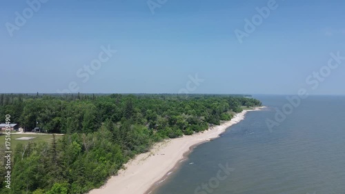 Сinematic drone footage follows the gentle curve of an untouched shoreline. The vibrant green of the forest contrasts with the pale sand and blue water, creating a peaceful and scenic natural landscap