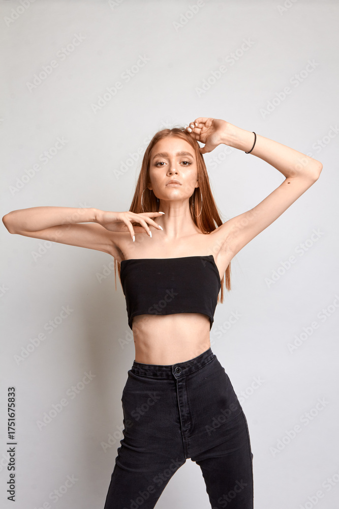 Naklejka premium A beautiful woman with a perfect figure is wearing black trousers and a black top. She stands on a white background in the studio.