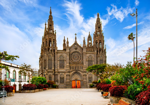 Front view of the Church of San Juan Bautista in Arucas, Gran Canaria, with ornate portal, palm trees, bushes, benches and large open square.