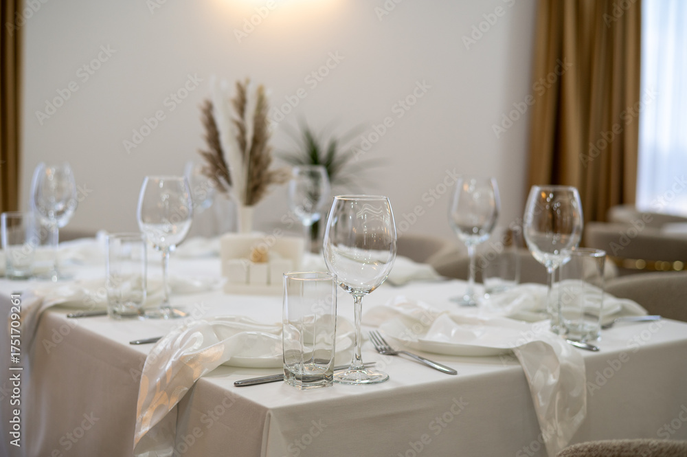 Fototapeta premium A sophisticated restaurant table arranged with white linens, crystal glassware, polished cutlery, and a delicate floral centerpiece. Plush seating and ambient lighting create an inviting, upscale
