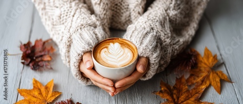 Cozy hands holding a latte with heart-shaped foam art surrounded by autumn leaves on wooden table in warm sweater