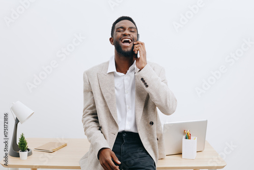 Confident young smiling man talking on mobile phone in modern office, casual business attire, cheerful expression, light background, workspace with laptop and greenery, professional lifestyle concept