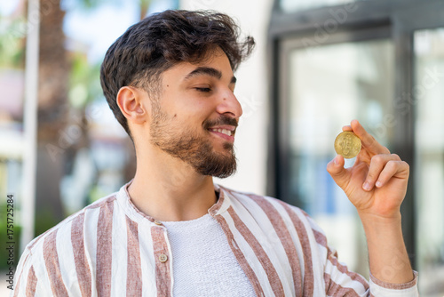 Handsome Arab man holding a Bitcoin at outdoors with happy expression