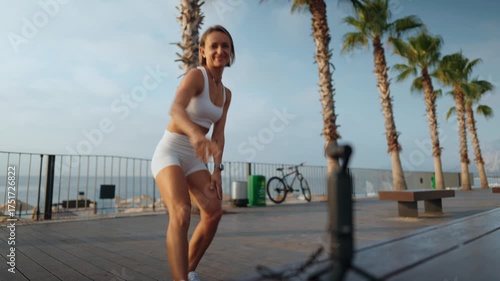 Fitness blogger woman films herself while walking outdoors on a sunny morning. She holds a camera in one hand and carries a yoga mat over her shoulder. Concept of active lifestyle and content creation