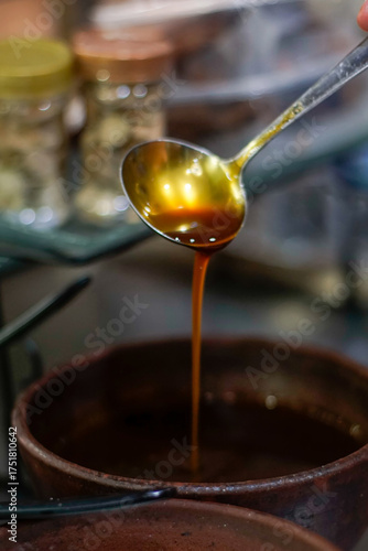 Close-up of liquid palm sugar (gula aren/kinca) being poured from a metal ladle into a rustic earthenware bowl. Warm amber syrup with glossy texture and artisanal kitchen background