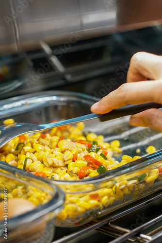 Close-up of stir-fried corn with sliced red and green chili being mixed with a spoon in a glass container, traditional Indonesian homemade dish.