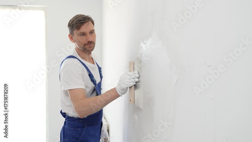 Fotografie Male construction worker wearing protective gloves and blue construction coveralls, spreading plaster smoothly across wall using professional drywall taping knife during renovation project