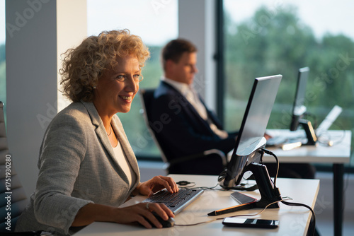 Smiling mid adult businesswoman working at her desk, typing on a keyboard and using a mouse while collaborating with a colleague in a bright, contemporary office environment