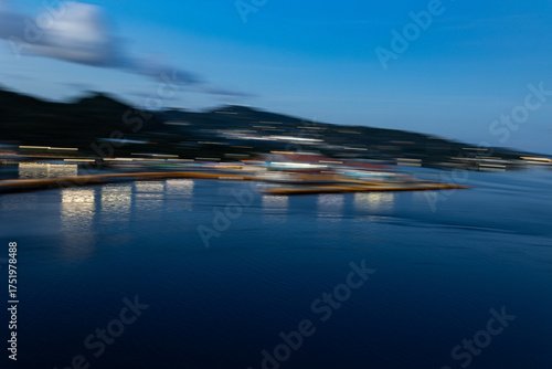 Abstract blurred night scene of a harbor and coastline with city lights reflecting on blue water.