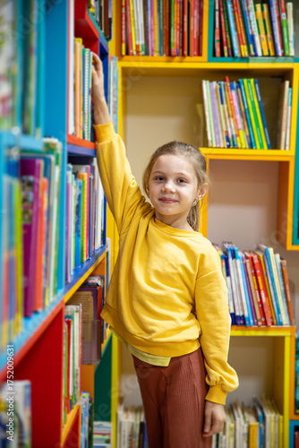 Portrait of a 7-8-year-old blonde girl in a yellow longsleeve in a library with books