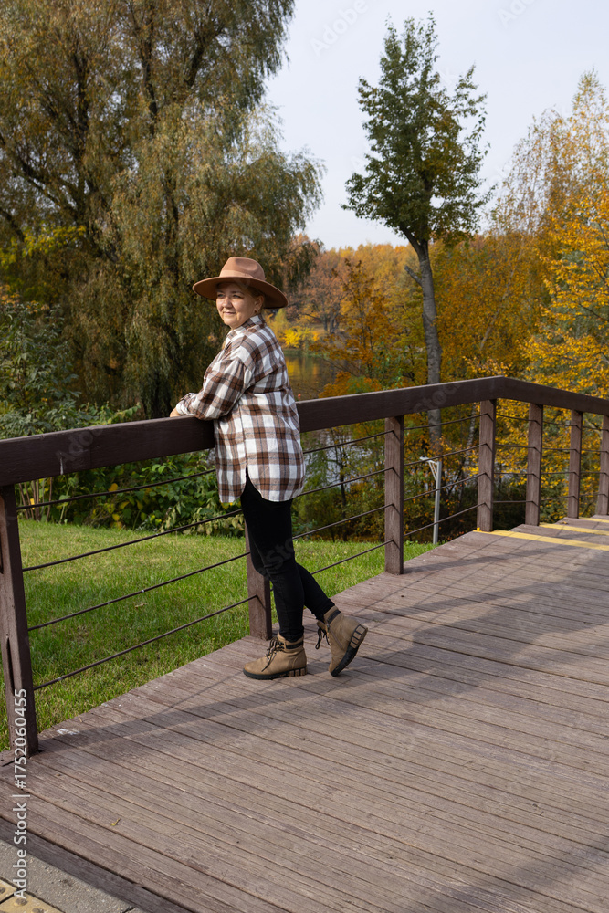 Fototapeta premium Caucasian woman with long hair stands on a on the wooden bridge surrounded by autumn trees. She wears a plaid shirt and a wide-brimmed hat.