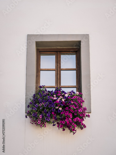 A rustic wooden window framed by a simple white wall, decorated with a vibrant arrangement of purple and pink flowers. The scene evokes charm and elegance in a minimal architectural style.