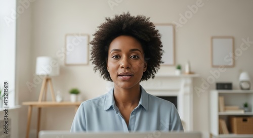 Professional woman speaking directly to camera during a virtual meeting from her bright home office, video call participant in a minimalist workspace with modern furniture and decor.