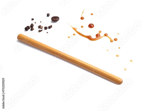 A light-colored wooden stirring stick is accompanied by scattered coffee grounds, a single bean, and a caramel-colored liquid splash on a white background