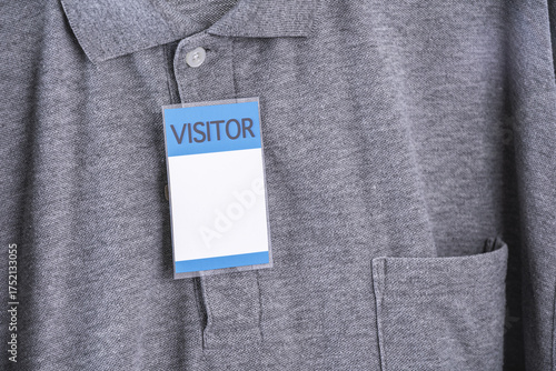 Visitor badge is attached to gray shirt, featuring collar and pocket. badge is prominently displayed, indicating temporary guest status