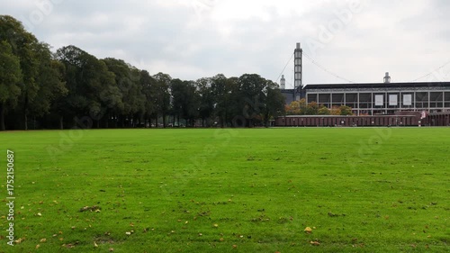 Cologne, Germany - 12 October, 2025: Wide lawn in front of a modern football stadium in Muengersdorf under cloudy sky. Long facade with brick pavilions and trees. Urban park sports venue. No people.