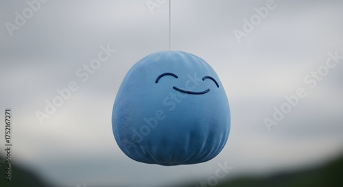 A blue plushie with a smiling face hangs from a string against a blurry, overcast backdrop