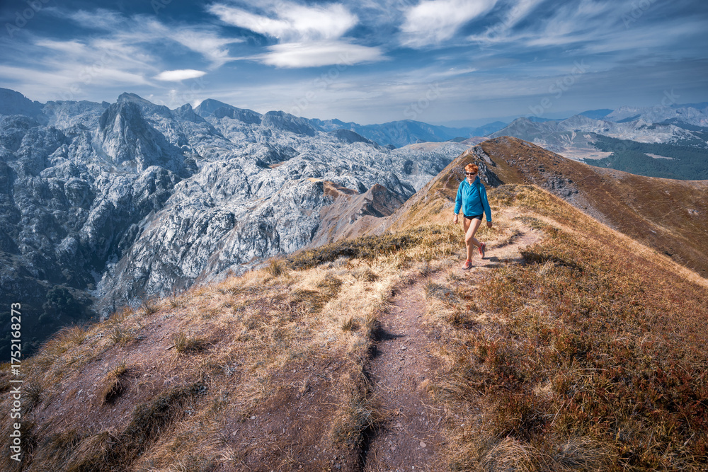 Fototapeta premium Female hiker walks narrow path to sunlit hilltop mountain ridge sunny day, Prokletije, Montenegro