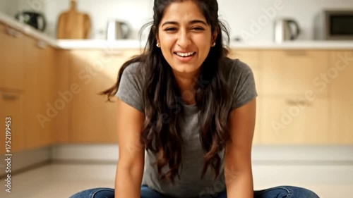 A cute Indian woman sits cross-legged on the kitchen floor surrounded by spilled rice, trying to pick it up grain by grain while laughing at her clumsiness