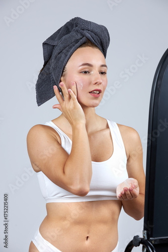 A young beautiful girl in white lingerie with a towel on her head stands in front of the mirror and does cosmetic procedures.