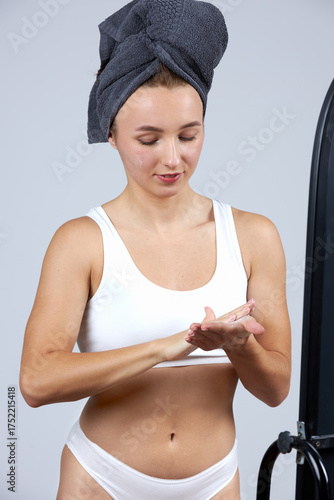 A young beautiful girl in white lingerie with a towel on her head stands in front of the mirror and does cosmetic procedures.