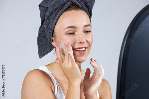 A young beautiful girl in white lingerie with a towel on her head stands in front of the mirror and does cosmetic procedures.