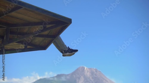 Person sitting on the edge of a wooden platform overlooking a snowy mountain peak under a clear blue sky.