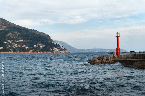 Red lighthouse on the pier in the port of Dubrovnik, Croatia, with the Adriatic Sea and coastal mountains in the background. Peaceful seascape under a cloudy sky
