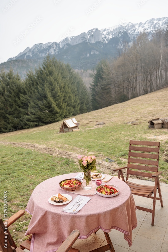 Fototapeta premium A round table with a pink tablecloth is set for lunch on a patio. Beyond the table, the landscape is a grassy hillside, pine trees, and the majestic Carpathian Mountains