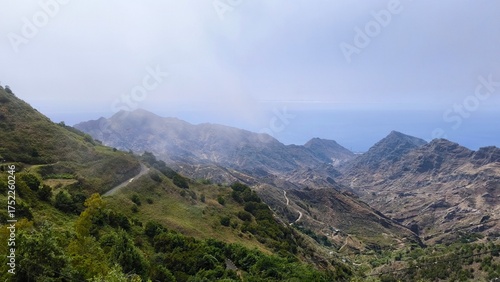 Mountain landscape of northern Tenerife, Canary Islands. View from TF-12 road with green slopes, winding paths, and the Atlantic Ocean in the distance under soft misty light.