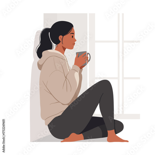 Woman sits by the window, holding a mug, lost in thought, wearing a hoodie and leggings.