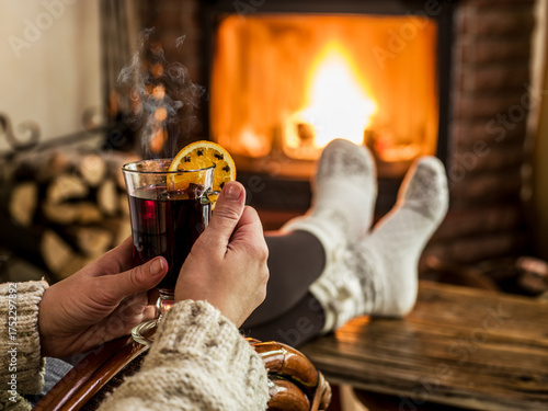 Glass of mulled wine or hot wine in woman hands and a burning fireplace in the background. Holiday background of warming up and relaxing at home.