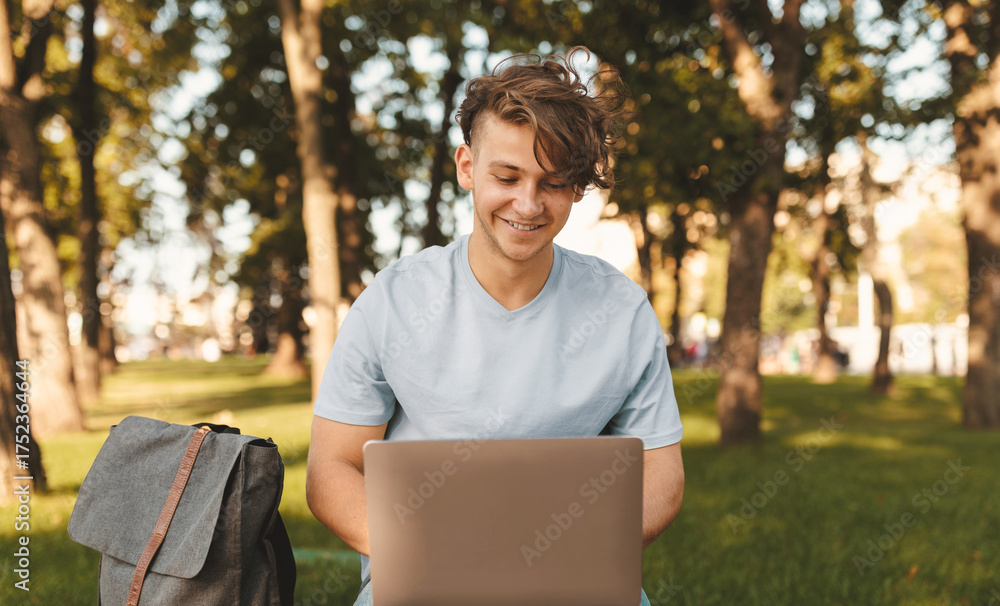 Fototapeta premium Cheerful student guy with laptop computer doing homework or attending online lecture, sitting on bench outdoors. Distance education concept