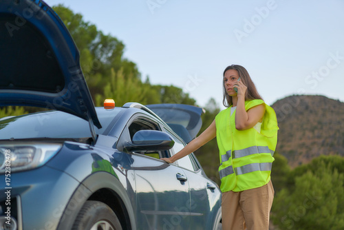 Girl in a yellow vest talks on her phone while her car is broken down on a rural road in the afternoon
